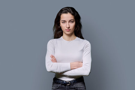 Beautiful young woman with crossed arms looking at camera on grey backgroundの写真素材