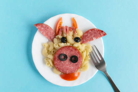 Funny Breakfast, lunch or dinner for children in the form of a bull's face made of pasta, sausage, bacon, olives and fresh tomatoes on a blue background. Plate with creative food art Breakfast for children with a fork. Top view.の写真素材