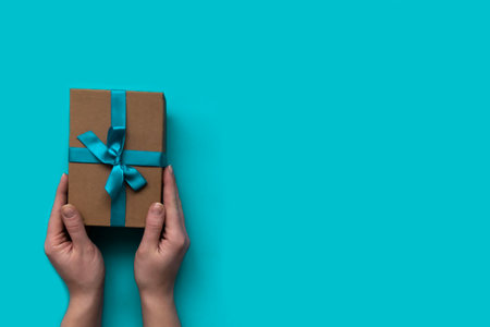 Female hands with a nice gift box decorated craft papper with a bow on blue background. New Year, Christmas, birthday, Valentines Day, World Womans Day, Fathers Day. copy space and top viewの写真素材