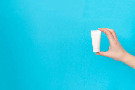 Womans hand holding white cream tube on a blue background. Beauty concept, clean and care cosmetics.の写真素材