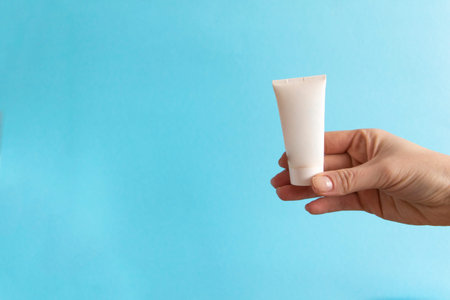 Womans hand holding white cream tube on a blue background. Beauty concept, clean and care cosmetics.