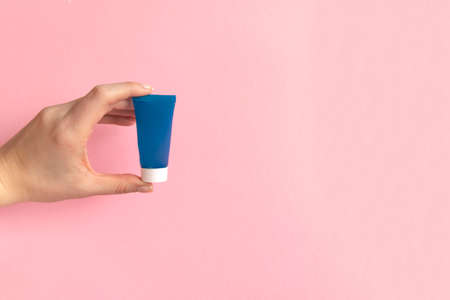 Womans hand holding blue cream tube on a pink background. Beauty concept, clean and care cosmetics.の写真素材