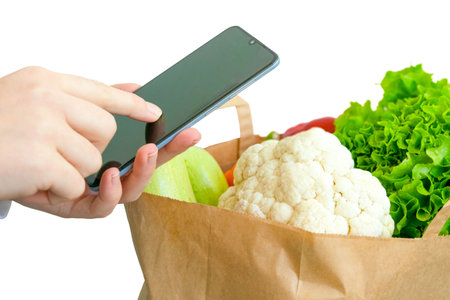 Smartphone in a womans hand. Fresh food vegetables donation isolated on background in paper bag package. Healthy food delivery during coronavirus.の写真素材