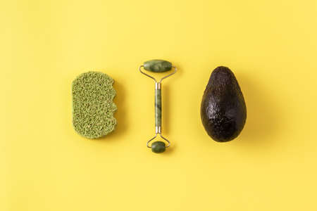 Jade roller, skincare tool for facial massage, bamboo sponge, avocado on yellow surface. Beauty treatment skincare routine concept. Plastic free beauty essentials. Flat lay, top view, creative layout.の写真素材