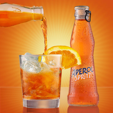 Bottle and glass of aperol spritz cocktail with orange slice. The bartender pours aperol spritz into glass filled with ice cubes. Liquor on orange background.のeditorial素材