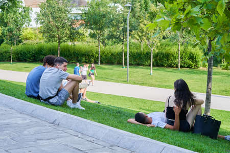 Teenagers hanging in the green park, relaxing, sitting and lying on grass. Boys looking on couple and talking to each other. Milan, Italy - June 16, 2020.のeditorial素材