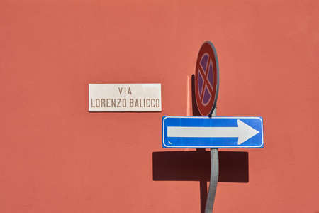 One way traffic sign, traffic road sign, sign with name of the street reading - Via Lorenzo Balicco - against brick colored wall - stop and parking are prohibited. Lecco, Italyの写真素材