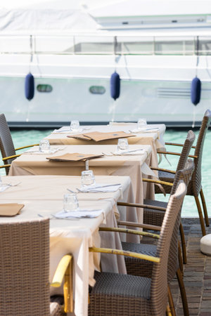 Outdoor dining tables set with glasses and napkins by a marina with yachts in the background, creating a luxurious dining atmosphere. Ideal for travel and hospitality themesの写真素材
