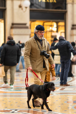 Milan, Italy - 18.12.2022: Man dressed in beige coat walks his black dog through in Gallery of Victor Emmanuel. Elegant setting and surrounding crowd create relaxed yet sophisticated city atmosphereのeditorial素材