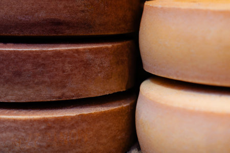 Close-up of stacked brown and yellow cheese wheels with textured rinds, showing slight imperfections and signs of aging, highlighting artisanal productionの写真素材