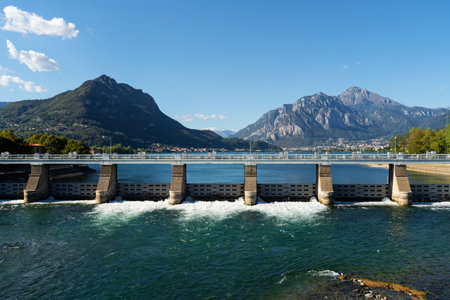 Large dam structure spans across lake with water flowing through the gates at lake Como, near Lecco, Italy. Towering mountains under clear blue sky with small boat visible on the lakeの写真素材