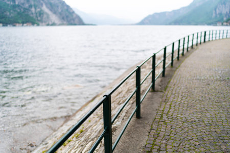 Scenic lakeside pathway with black metal railings, overlooking calm waters and distant mountains on cloudy day.の写真素材