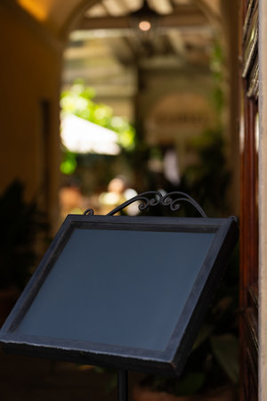 Close-up of blank blackboard sign with ornate frame in cozy outdoor cafe setting, featuring blurred greenery and sunlightの写真素材