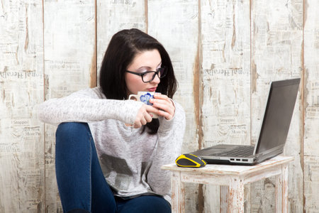 Student girl is wearing pair of glasses is studying on laptop while drinking tea from her mug.の写真素材