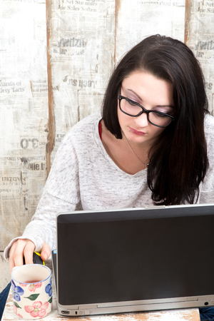 Student girl is wearing pair of glasses is studying on laptop while drinking tea from her mug.の写真素材