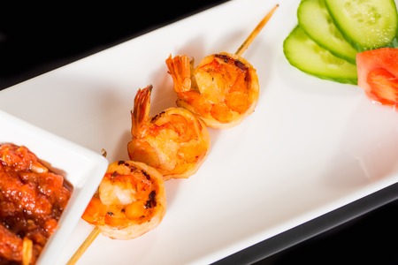 Fried kebab of shrimps with vegetables and sauce on white plate against black backgroundの写真素材