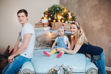 Dad and mom kissing little daughter from sides, posing in decorated for Christmas studio.