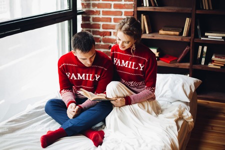 Couple in red sweaters reading book in bed together.の写真素材