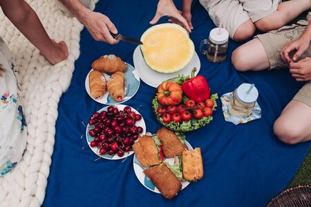 Family having picnic on the blanket. homemade croissants and sandwiches with lemonade. Man is cutting a yellow watermelon.の写真素材