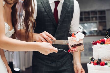 Side view of young bride and groom keeping knife and plate and cutting delicious wedding cake in restaurant. Piece of yummy sweet dessert. Concept of confectionery, celebration and love.の写真素材