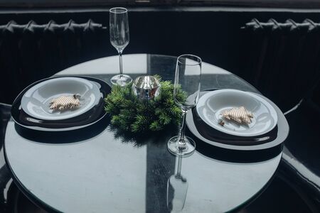 Close-up of round table served for two for Christms dinner. Silver candle decorated with natural fir branches, two flutes, pairs of plates with decorative handmade Christmas trees.の写真素材