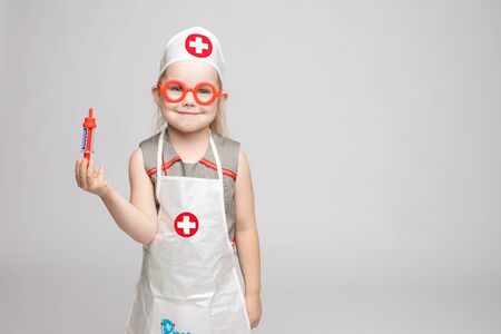 Little cute funny girl playing wearing doctor uniform holding toy syringe looking at cameraの写真素材