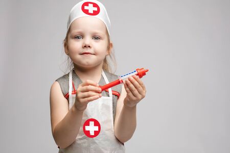 Little cute funny girl playing wearing doctor uniform holding toy syringe looking at cameraの写真素材