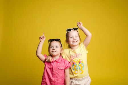 Two happy beautiful stylish little girl wearing sunglasses posing isolated yellow studio backgroundの写真素材