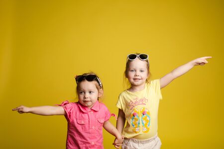 Two happy beautiful stylish little girl wearing sunglasses posing isolated yellow studio backgroundの写真素材