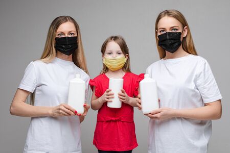 Portrait of three handsome girls with long fair hair with medical masks on their faces holds a lot of antisepticの写真素材