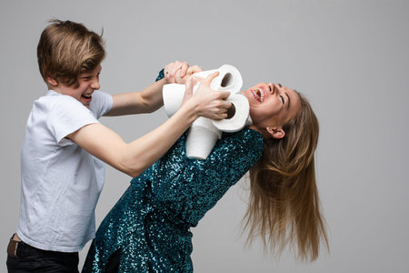 Young cheerful woman fights for a lot of toilet paper with a young boy, portrait isolated on white...