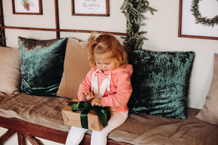 Little pretty girl sith wavy hair sits of the sofa opens a box with a present