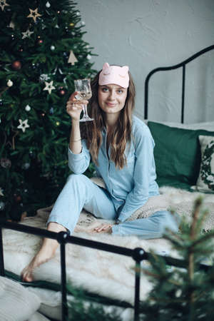 Beautiful woman with drink on bed. Smiling jolly woman in blue pajamas and sleeping mask sitting on bed with glass of champagneの写真素材