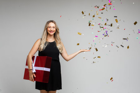 Smiling young girl with large festive gift pointing by hand on empty space with holiday confetti on...