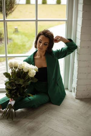 portrait of beautiful female in green suit poses for the camera with a lot of white roses in her hands near the big windowの写真素材