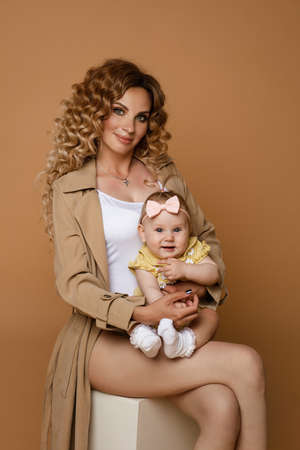 Gorgeous woman in beige trench with a baby daughter and white top holding cute baby daughter in arms sitting on white against peach background.の写真素材
