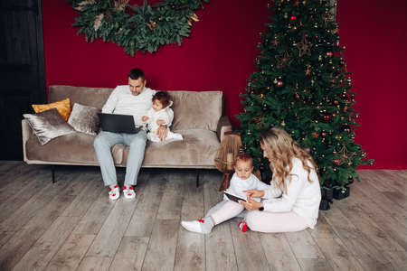 Dada and son sits on a sofa together, mum and daughter play near the christmas treeの写真素材