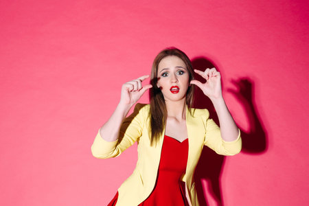 Pretty fashionable girl in red dress and yello jacket posing on pink background.