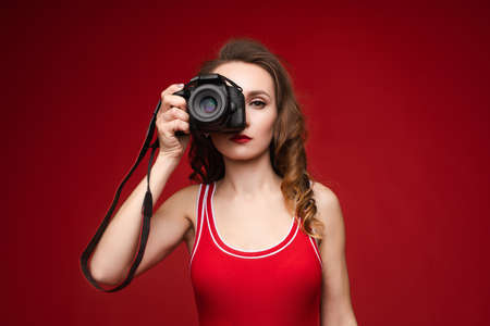 A beautiful bright woman with a camera in her hands. A woman on a red background in a red swimsuit poses with a camera.の写真素材