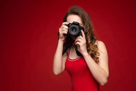 A beautiful bright woman with a camera in her hands. A woman on a red background in a red swimsuit poses with a camera.の写真素材
