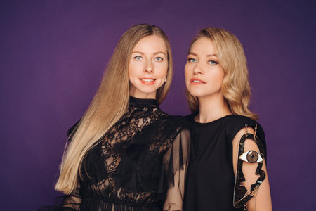 Two sisters pose in the studio on a purple background. 2 beautiful women in black fancy dresses gathered for a party.の写真素材
