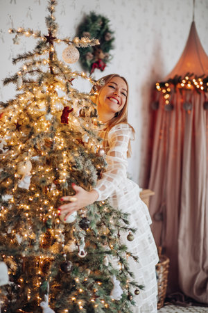 A beautiful stylish woman in a white dress near the Christmas tree is smiling joyfully and wishes happinessの写真素材
