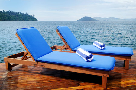 Pair of outdoor reclining seats on deck near large lake with hills in the distance. Matching blue and white towels in rolls on top.の写真素材