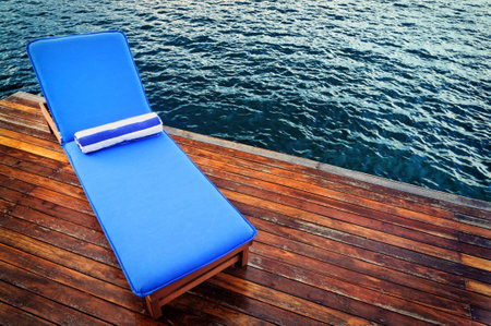 Outdoor reclining seat on deck next to lake. Matching blue and white towel rolled up on top.の写真素材