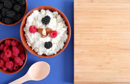 Cottage cheese in a clay bowl with berries on a blue background top view. Fermented milk product with raspberries and blackberries on blue with space for text.の写真素材