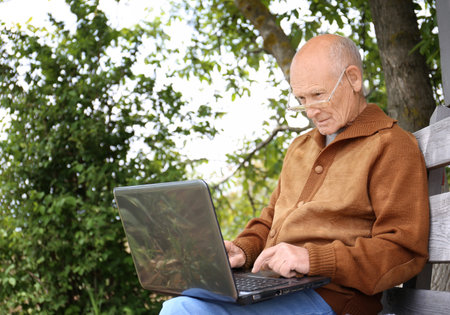 An old man sits in nature and studies a laptop. Grandfather on a bench studying a laptop. Old man...
