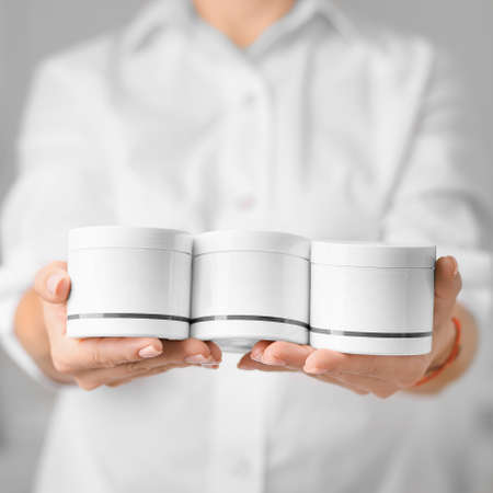 Hands of young woman holds hand skincare cream. Woman model with body cream jar in hands. Winter skin care and wellness concept. Close-up closed jars of cream in hands. The beautician holds products in the form of jars of cream.の写真素材