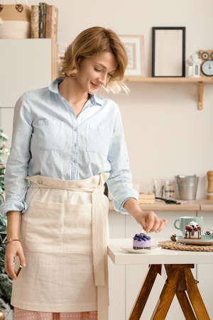 A young girl in the kitchen lights a candle for a cupcake for her birthday. A lonely girl makes a holiday for herself. Happy girl in an apron lights a candle in the kitchenの写真素材