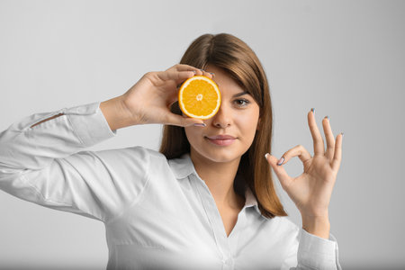 A girl with an orange advertises a cream. Healthy skin. A girl in a white shirt closes her eye with an orange and shows ok on a gray backgroundの写真素材
