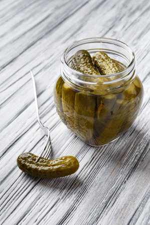 Pickled gherkins in an open glass jar and one cucumber on a fork nearby on a light wooden background close-up. Homemade pickled cucumbers.の写真素材